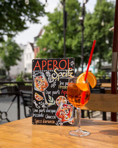 Aperol-Blechschild mit italienischem Rezept in Außenszenerie neben Glas Aperol Spritz