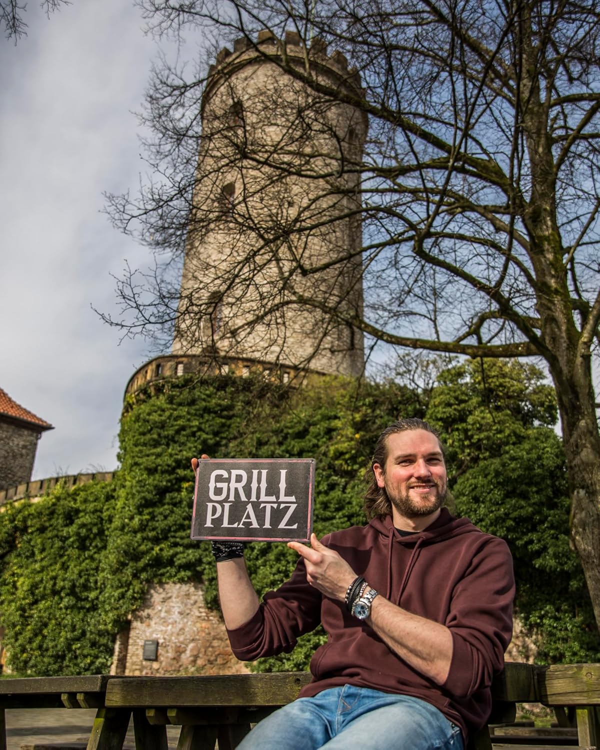 Mann sitzt vor Sparrenburg und hält Blechschild mit Aufschrift Grillplatz hoch