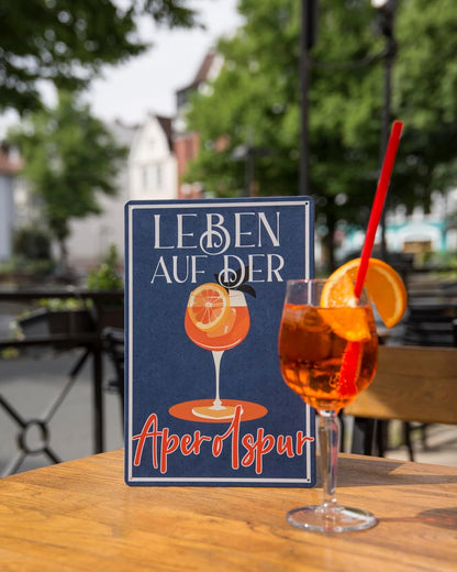 Dekoratives Blechschild mit Aufschrift Leben auf der Aperolspur mit Cocktail auf einem Tisch