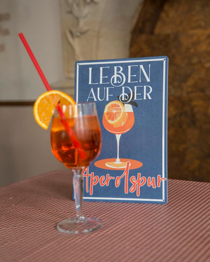 Blechschild mit Aufschrift Leben auf der Aperolspur mit Cocktailglas dekorativ auf einem Tisch